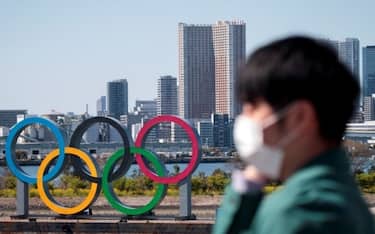 A man wearing a face mask, amid concerns over the spread of the COVID-19 novel coronavirus, stands before the Olympic rings from an observation point in Tokyo's Odaiba district on March 25, 2020, the day after the historic decision to postpone the 2020 Tokyo Olympic Games. - Japan on March 25 started the unprecedented task of reorganising the Tokyo Olympics after the historic decision to postpone the world's biggest sporting event due to the COVID-19 coronavirus pandemic that has locked down one third of the planet. (Photo by Behrouz MEHRI / AFP) (Photo by BEHROUZ MEHRI/AFP via Getty Images)
